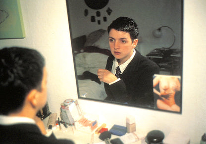Interior image - NAN GOLDIN / COUPLES AND LONELINESS. Person looking into a mirror in a room with various items on a table.